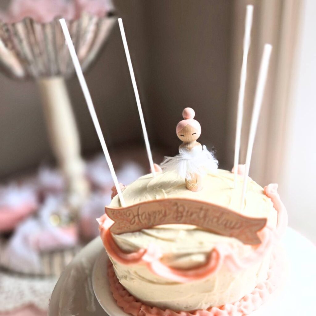 Small birthday cake with white and pink frosting and a wooden peg doll ballerina on top.